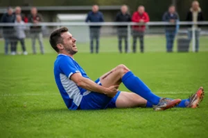 Joueur de football blessé à la jambe, allongé sur le terrain, souffrant, en tenue bleue et blanche.