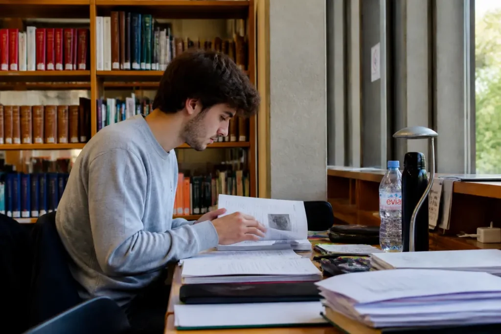Homme étudiant à une table de bibliothèque, entouré de livres et de cahiers, concentré sur son travail.
