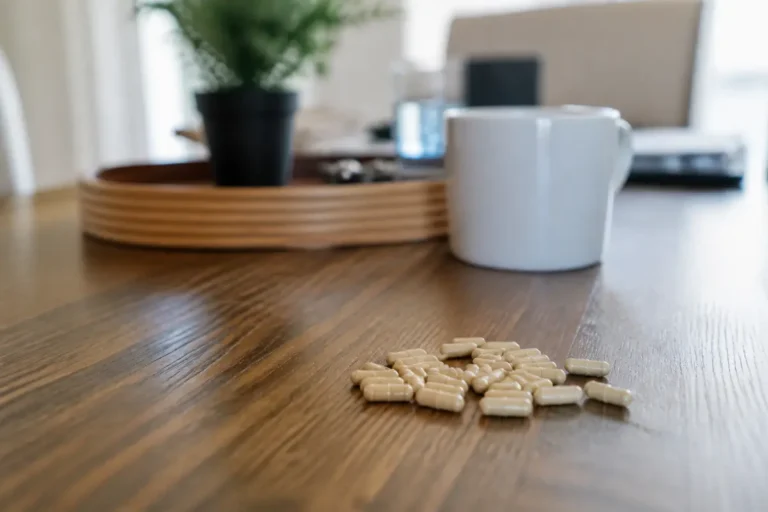 Capsules sur table en bois, avec tasse blanche floue en arrière-plan, et plante.