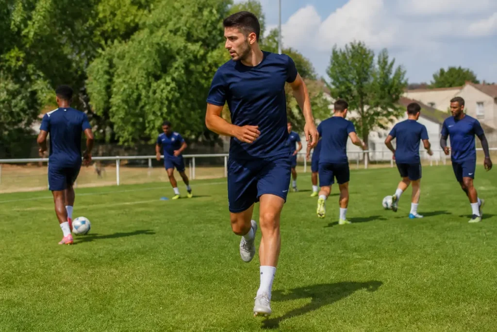 Joueurs de football en bleu s'entraînant sur un terrain herbeux, préparant un match sous ciel ensoleillé.