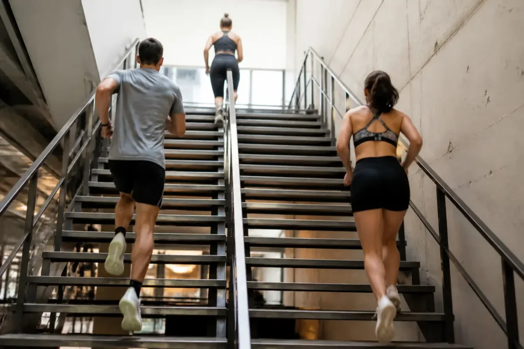 Personnes montant un escalier métallique en tenue de sport, intérieur moderne, efforts physiques visibles.