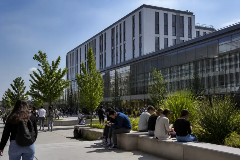 Des personnes assises sur un muret devant un bâtiment moderne, entouré d'arbres, par une journée ensoleillée.