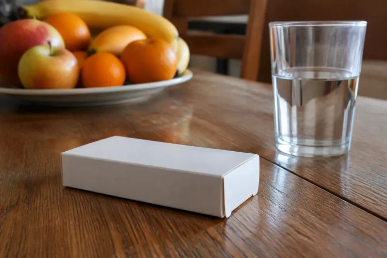 Verre d'eau, boîte blanche, assiette de fruits variés sur une table en bois.