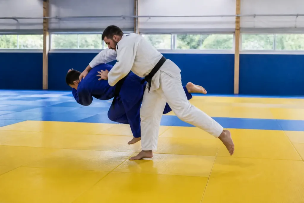 Deux judokas s'entraînent au judo sur un tatami jaune et bleu dans un dojo lumineux.