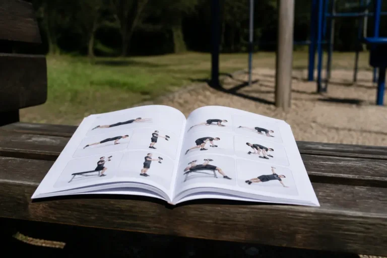 Livre d'exercices de fitness ouvert sur un banc en bois, parc en arrière-plan flou.