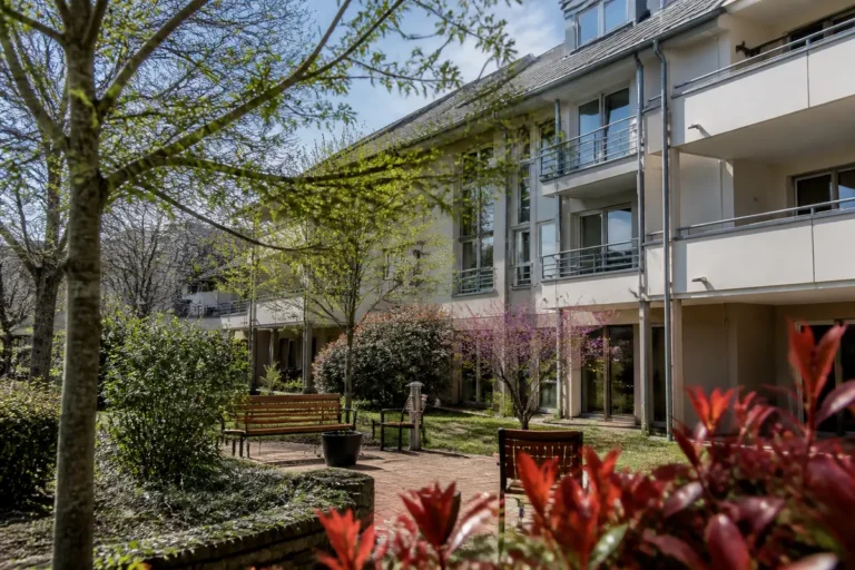 Cour arborée d'une résidence avec bancs, fleurs rouges et bâtiment moderne à trois étages en arrière-plan.